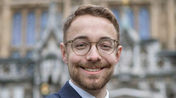 A man pictured in front of the House of Commons. He is smiling, has brown framed glasses and a brown beard