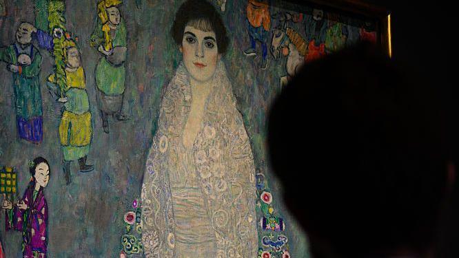 A man stands in front of a full-length Art Nouveau portrait of a woman.