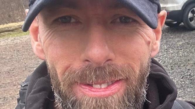 John Hall has a greying beard wearing a NY navy baseball cap. He is grinning and his teeth are visible in the very close up shot.