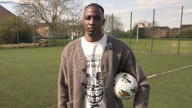 Joe Aribo stands with a football tucked under his left arm and looks directly into the camera while wearing a fuzzy fawn-coloured cardigan and white t-shirt with a black cartoon face on it. Aribo is standing on an astro turf pitch surrounded by a high green chain-link fence. Beyond the fence a park can be seen with blocks of flats beyond that