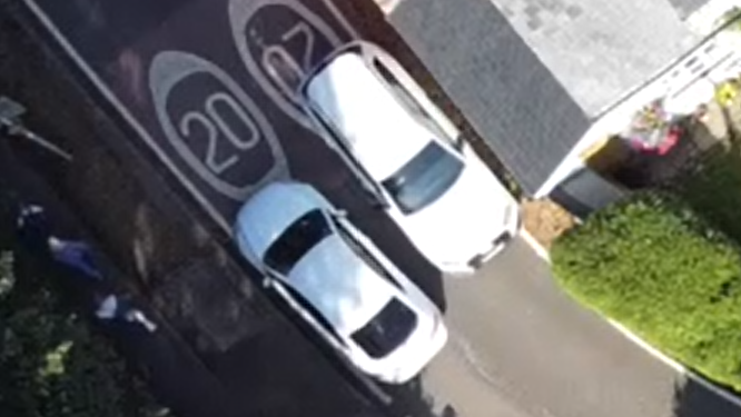 This is a drone shot of Walton Road in Wavendon village. You can see two white cars trying to pass each other from above and there isn't much of a gap between them. You can also see the roof of a house from above and two 20 mile an hour painted signs on the road.