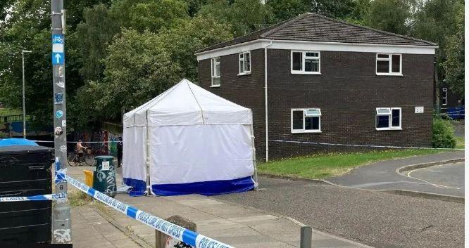 A police white forensic tent in a street cordoned with blue and white police tape. There are tall trees in the background.