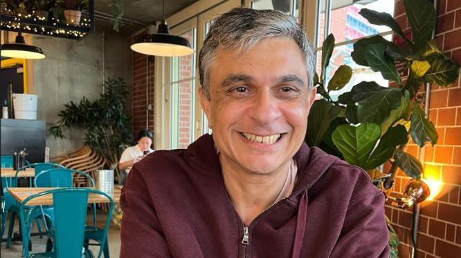 Close up image of Niraj Haria wearing a red zip-up top sitting in a cafe with chairs and table in background. He is smiling