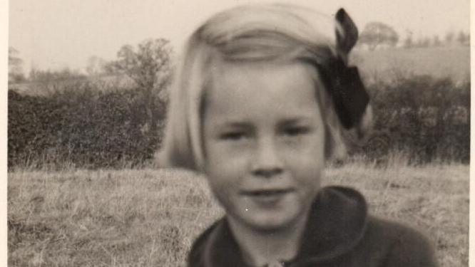 A sepia photograph of a little girl with short blonde hair tied at the side with a ribbon and a dark coat with a curved collar. Fields, trees and hedgerows are behind her.