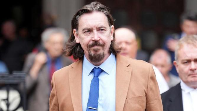 Richard Hall walks out of the court, wearing a light brown suit with a pale blue shirt and a darker blue tie. He has a beard and shoulder length brown hair. Mainly blurred images of other people can be seen in the background