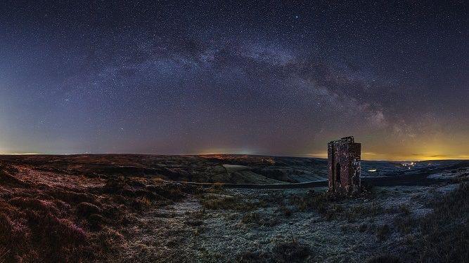 Dark skies above Rosedale, in the North York Moors. A landscape image, of moorland with the ruins of a tower to the left. Clusters of stars can be seen in the sky, which is a purple hue.
