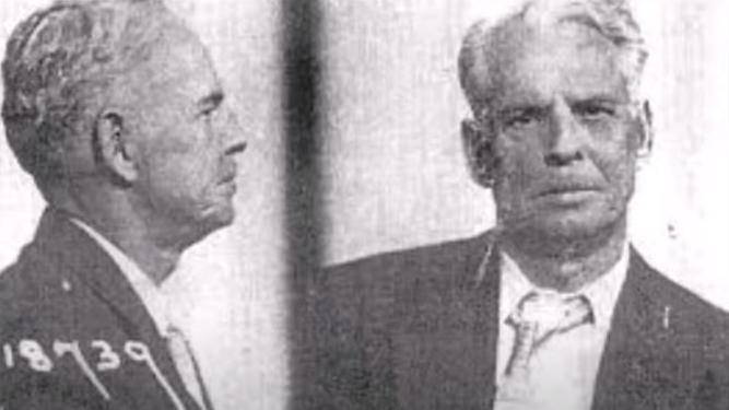 An FBI mugshot from the day of his arrest on 23rd November 1965. He has white wispy hair, and his tie and shirt look dishevelled