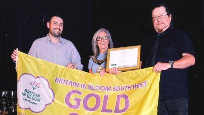 Representatives from Yeovil hold a large yellow banner with purple writing, which reads, "Britain in Bloom South West, Gold 2025. One of the three holding the banner, a woman, also displays a certificate of the award in her left hand. All are smiling at the camera.