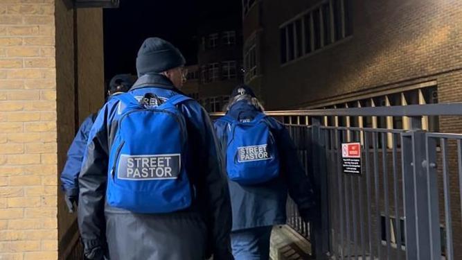 Three people with their backs to the camera are dressed in blue uniform, with backpacks that read "street pastor". They are outside, with a brick wall on their left and a metal railing on their right.