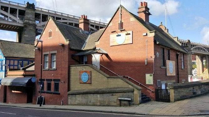 A picture from 2015 showing a brickbuilt building just next to a bridge in Newcastle with a stone staircase and a sign saying River Beat 