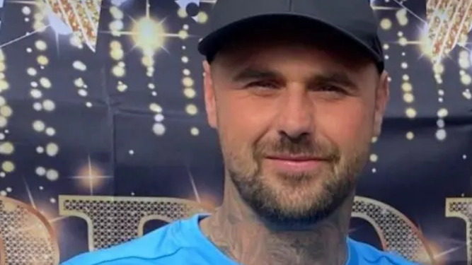 Tom Gaughan has a neatly trimmed, dark beard and tattoos on his neck. He is wearing a blue top and a North Face cap and is smiling into the camera.