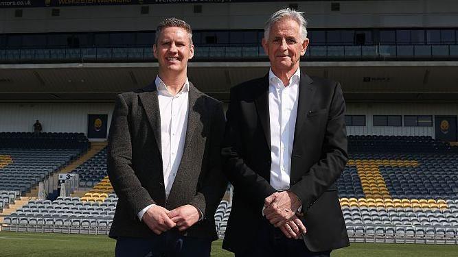 Stephen Vaughan stands next to Worcester Warriors owner Chris Holland in font of the main stand at Sixways. Both are wearing dark suits with white shirts and have their hands crossed in front of their waists. 