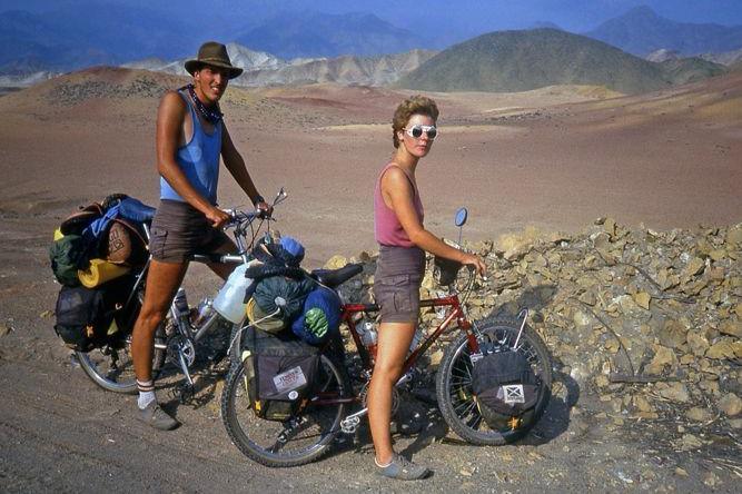 Craig and Rona pose for a picture on their bikes with a desert and rugged mountains behind them. Craig is wearing a wide brimmed hat, a blue vest and grey shorts. Rona has stylish white-rimmed sunglasses, a pink vest top and grey shorts.