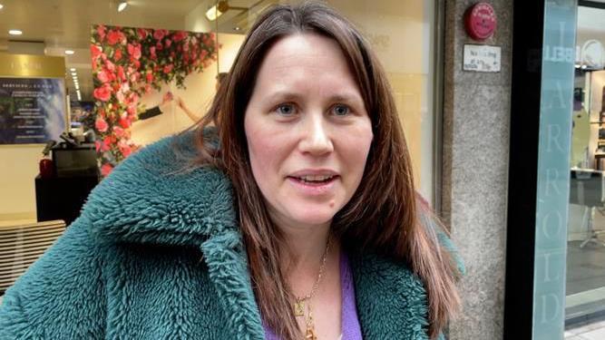 Rachel Martin, a woman looking at the camera and smiling. She has brown hair and is wearing a blue fluffy coat and purple top with gold necklaces. She is stood in front of a shop window. 