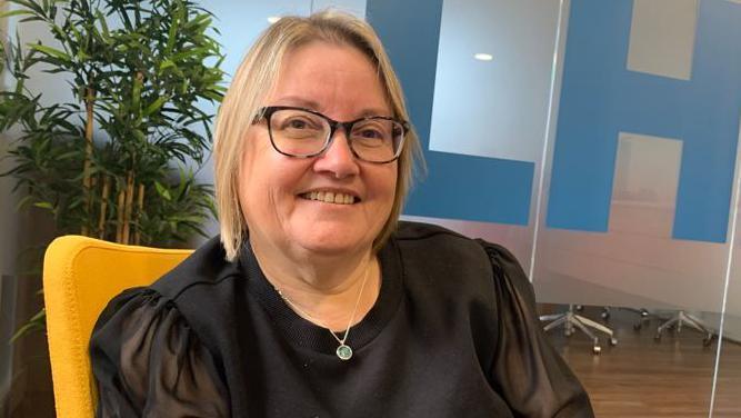 Woman in a black top and glasses sitting down and smiling at the camera. She has short blonde hair. 