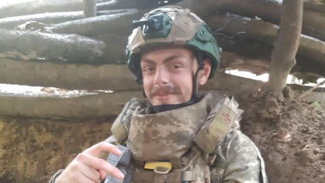 Callum Tindal-Draper wearing military camouflage clothing while stood in front of a set of logs in Ukraine.