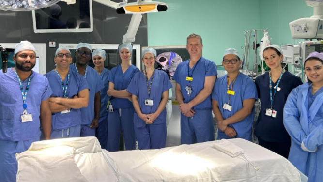 A group of men and women line up in a medical room infront of a hospital bed. They are all dressed in blue scrubs.