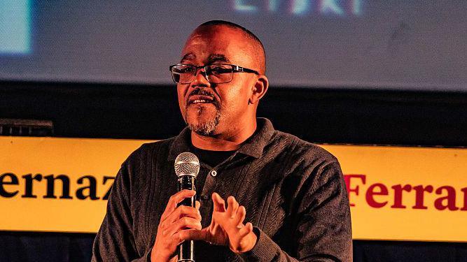 Kehinde Andrews, a black man with a dark shirt, square-framed glasses and a black and grey beard, stands on stage holding a microphone.