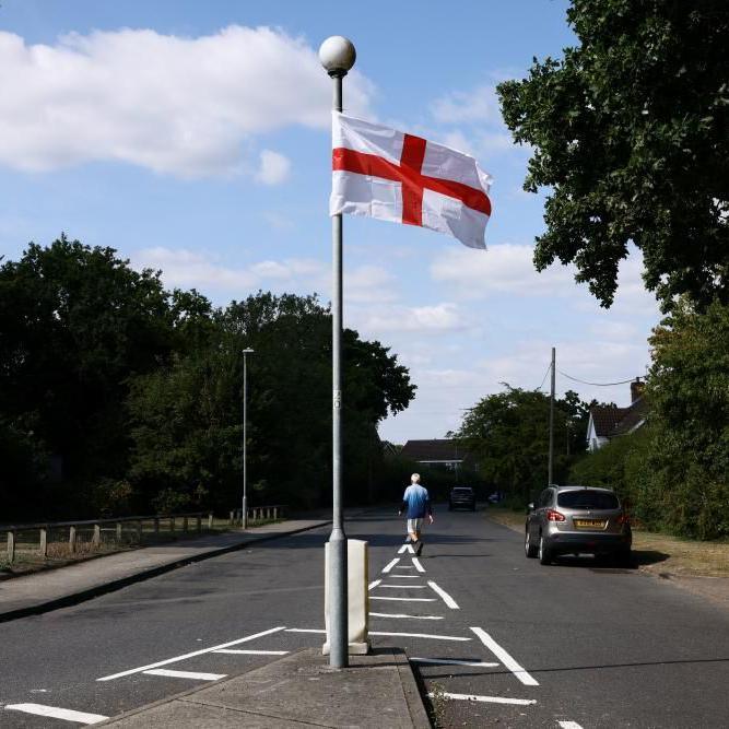 A flag of St George hangs from a lamppost on an island in the middle of an empty road, as a man walks down the road in the background.