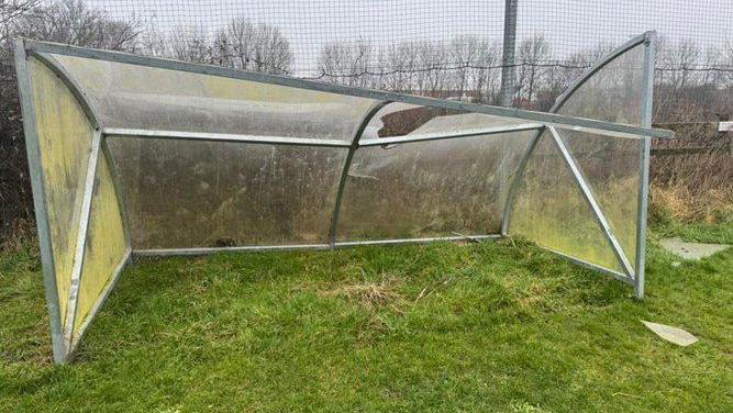 A broken transparent plastic shelter with a metal frame stands on grass, its roof collapsed and a side panel bent inward. A wire fence and bare trees are visible in the background beneath a grey, overcast sky.