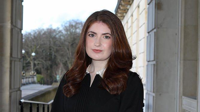 Connie Egan, a woman with long, brown, wavy hair, looks directly at the camera while standing outside Parliament Buildings at Stormont. She is wearing black dress with a white collar