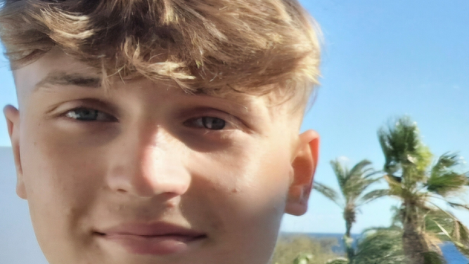 Harvey Nye, aged 19 and from Plymouth, stands in front of a pair of palm trees on a sunny day with the sea in the distance. He is smiling and the fringe of his mousey coloured hair is covering most of his forehead.