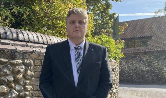 Gregory Hill, wearing a dark suit, white shirt, and a grey, black and white striped tie, stands solemn-faced against a flint wall and in front of a road and house.