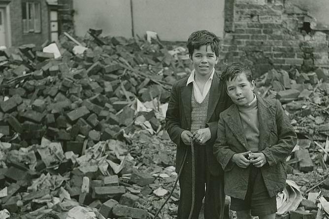 Two children stood in front of a collapsed building with a pile of bricks next to a damaged wall. They wear coats over jumpers and one appears to hold a length of rope.