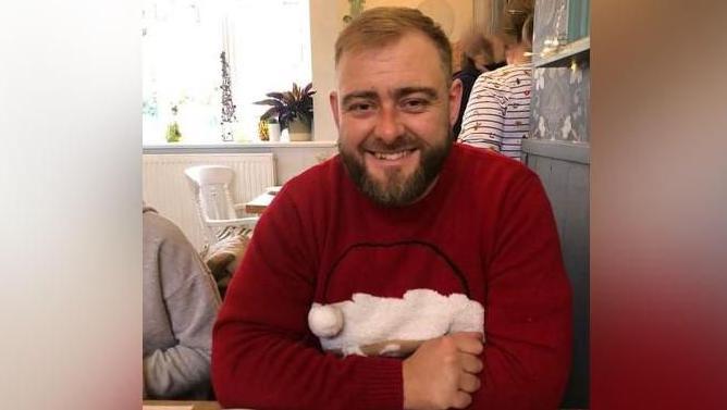 Andrew Darn pictured smiling and sitting at a table. He is wearing a red jumper with a Santa Claus on it.