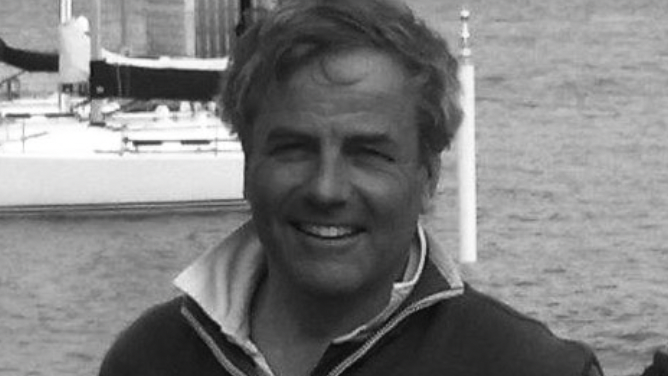 Headshot of Robin. He is smiling at the camera and stands on the coast - with the sea and a few boats behind him.