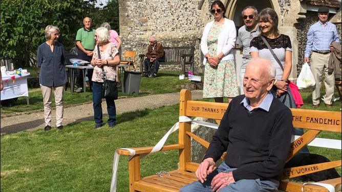 An elderly man is sitting on a bench in front of a church. A group of people behind him are smiling. It's a sunny day.