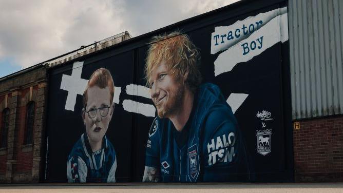 An Ed Sheeran mural painted on the side of a building. A child version of Sheeran as well as how he looks now are painted side by the side. They both wear Ipswich Town shirts.