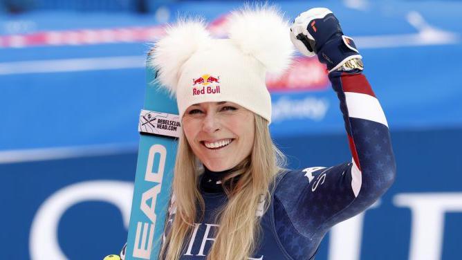 Lindsey Vonn is wearing a white hat with furry ears and holds up her arm