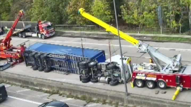 Cranes lifting a blue and white lorry back onto its wheels. It is on its left side across a four-lane motorway. Two mobile cranes are parked at the front and back of the lorry