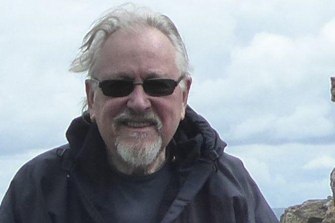 Ian has grey hair which is blowing in the wind. He is wearing sunglasses and has a small goatee grey beard. He is wearing a dark walking jacket.