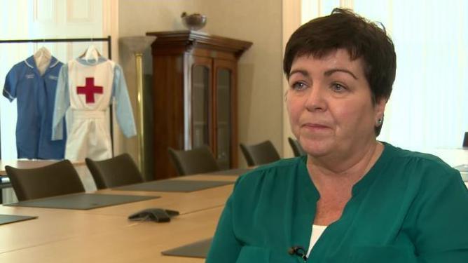 An image of Rita Devlin from a previous BBC TV interview. Ms Devlin has short, dark brown hair and is wearing a jade green blouse over a white top. There is a table and glass-fronted cupboard in the room behind her. Two nurses uniforms are hanging on a rail by a window