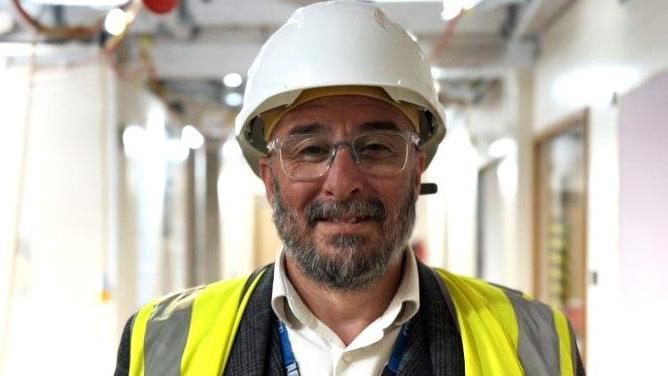 Stephen Balls with beard and glasses wears a hard hat and high-visibility vest. He stands in a hospital corridor where construction work is being carried out.