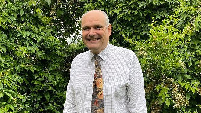 Michael de Whalley the candidate for North West Norfolk standing in front of a green hedge, wearing a shirt and floral tie 