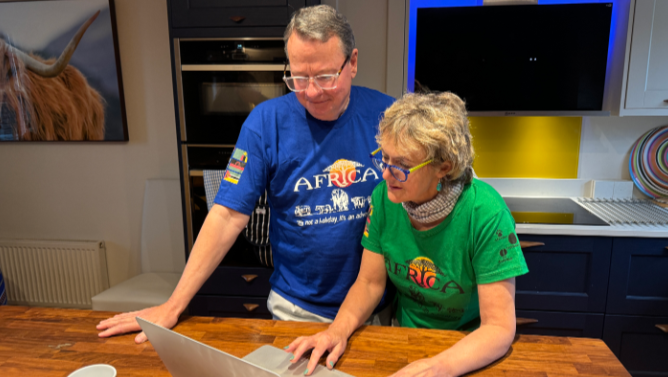 The couple at home in their kitchen. Mr Cherry is wearing a blue T'shirt and his wife has a green T'shirt on. They are both looking at a laptop on their table.