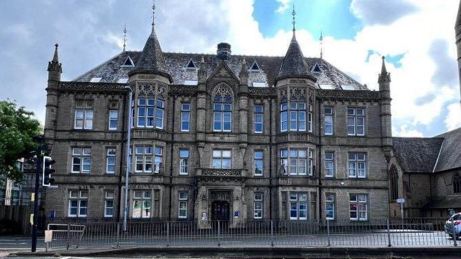 A large Victorian building with three storeys, turrets and many windows. It is set back from what appears to be a main road. A traffic light and pedestrian railings are visible in the foreground.