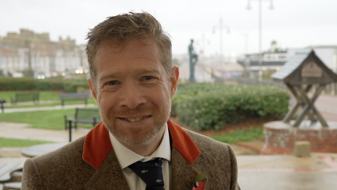 A head and shoulders image of Zeb Soanes smiling while looking into the camera. He is wearing a tan and orange blazer over a white shirt and navy tie. He is standing outside and it is raining.