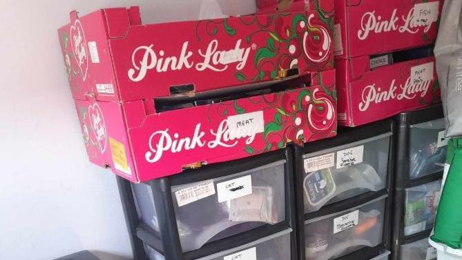 Containers and cardboard boxes in Wendy Isaacs' spare room, where she stores supplies for the pet food bank. Four Pink Lady apple boxes are stacked near the wall, labelled with their contents. Drawers can be seen below them, also labelled with the contents. 