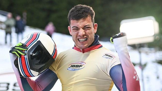Matt Weston celebrates. He has short dark hair and is looking directly into the camera and raising his left fist. He is holding his crash helmet in his other hand.
