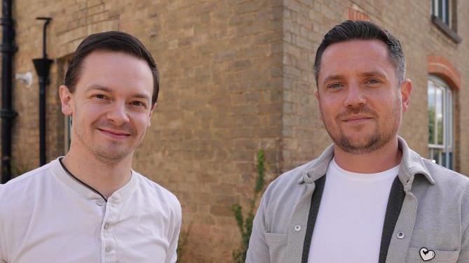 Chris Tuczemskyi and Ben Kennedy. Both are looking into the camera. Chris is wearing a collarless white, long sleeved shirt and Ben is wearing a white t-shirt and light grey jacket. They are standing on the corner of a brick building. 