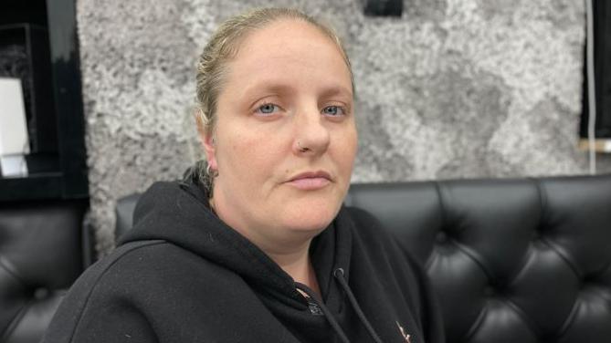 Zoe Savva, with a serious expression, is wearing a black hoodie and has her blonde hair pulled back, and is sitting on a black sofa.