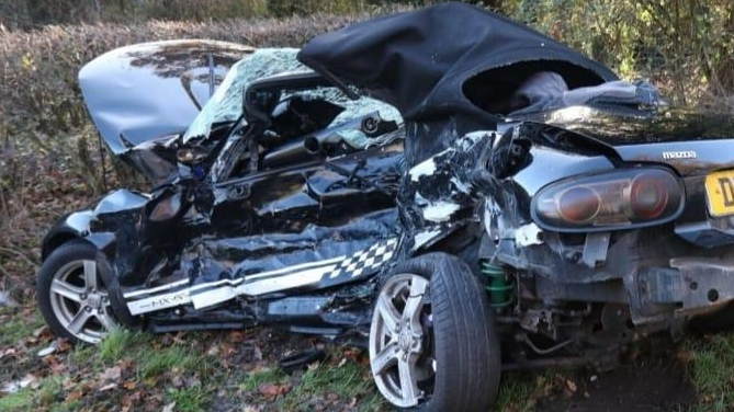 A very badly damaged car, with a smashed windscreen, the passenger side bent inwards and the rear tyre off the wheel. The car is pointing into a hedge