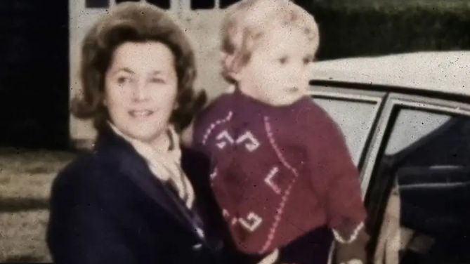 Renee MacRae holds toddler Andrew in her arms in a picture taken in the 1970s. Mrs MacRae is wearing a dark jacket and Andrew wears a red jumper with a white pattern on it. They are next to a car.
