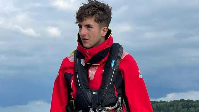 Image of Thomas Roy he is wearing red waterproofs and a black life vest at the helm of a boat