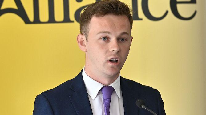 A man speaks into a microphone. He has short brown hair and is wearing a navy suit, white shirt and purple tie. He is standing in front of a yellow background with the word 'Alliance' written in large black letters.