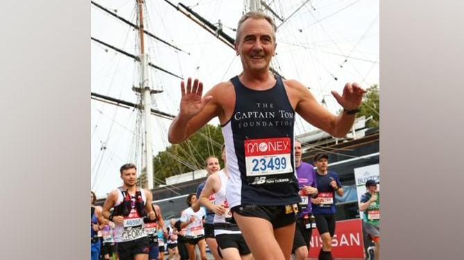 Liam Mannion   running the London Marathon in 2021 for the Captain Tom Foundation. He is wearing a navy and white vest with the charity's name on it and there are other runners behind him as well as the mast and ropes of a ship.